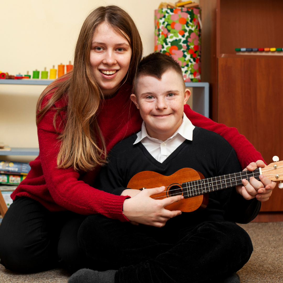 A boy playing a small guitar smiles at the camera with a woman sat with her arms round helping him