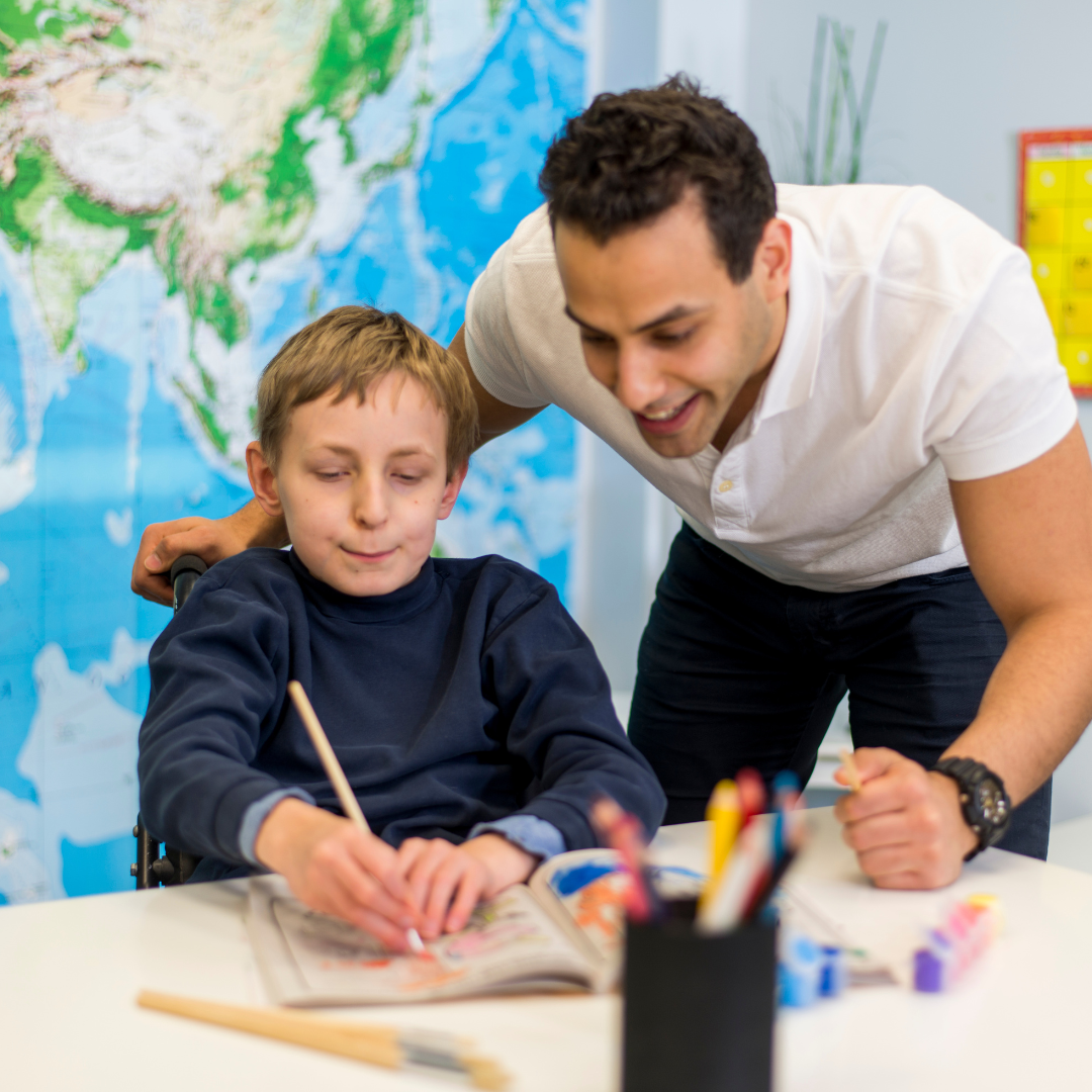 A boy sat at a table in a classroom drawing and a man leaning over him smiling