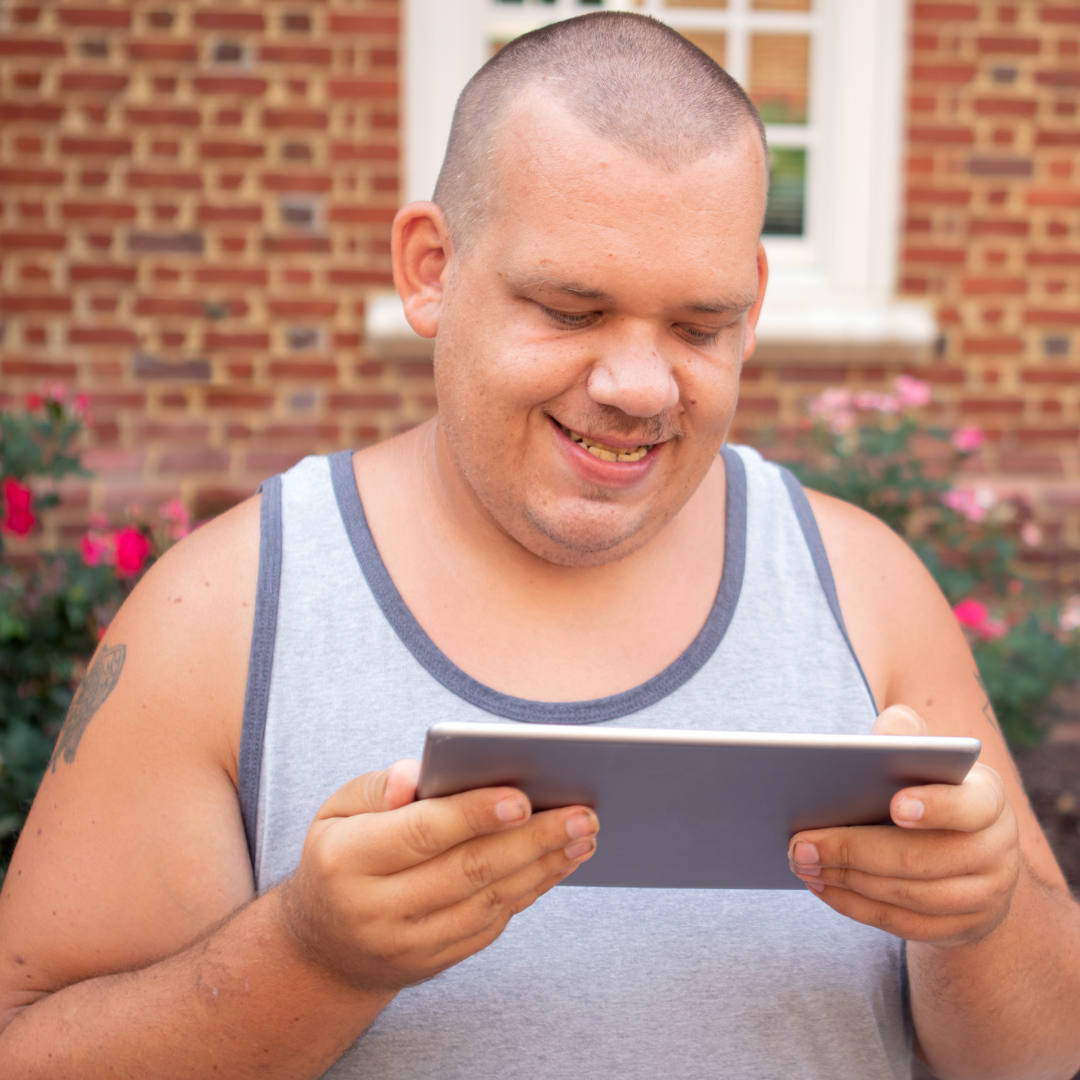 A man with a shaved head wearing a vest looks down at a tablet device smiling