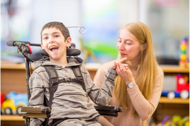 A boy in a wheelchair in a school setting with a female adult crouched down beside him