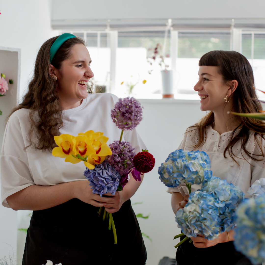 Tw ofemales holding bunches of flowers in a flower shop look at each other smiling
