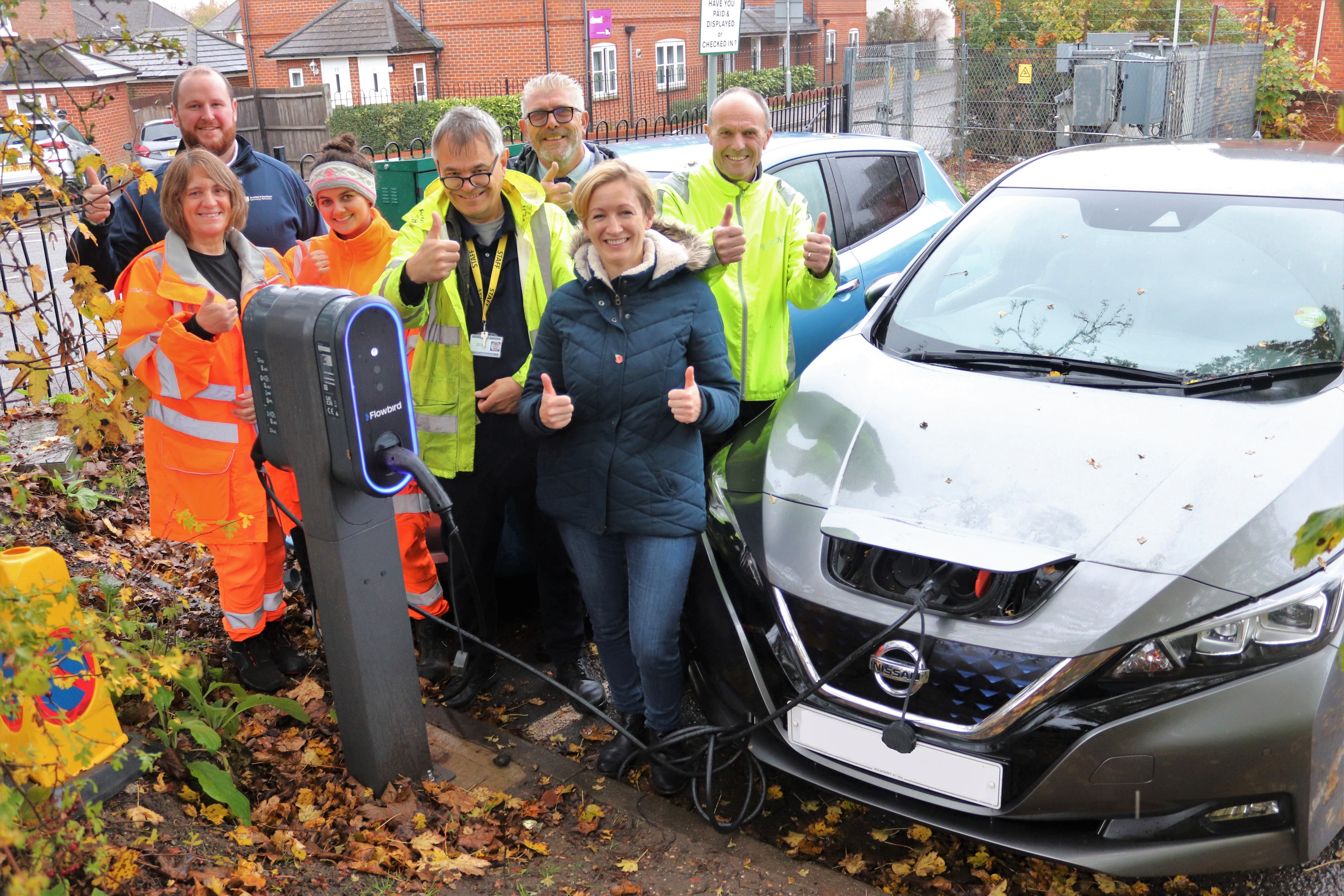 A group of people in mostly high viz jackets gather around an electric vehicle as it charges in a car park