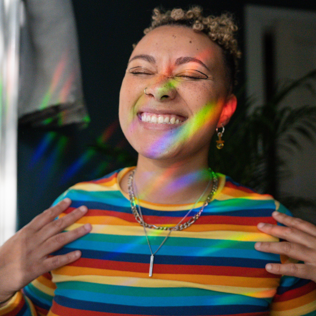 A person wearing a rainbow t-shirt with a rainbow reflection across their face closes their eyes smiling