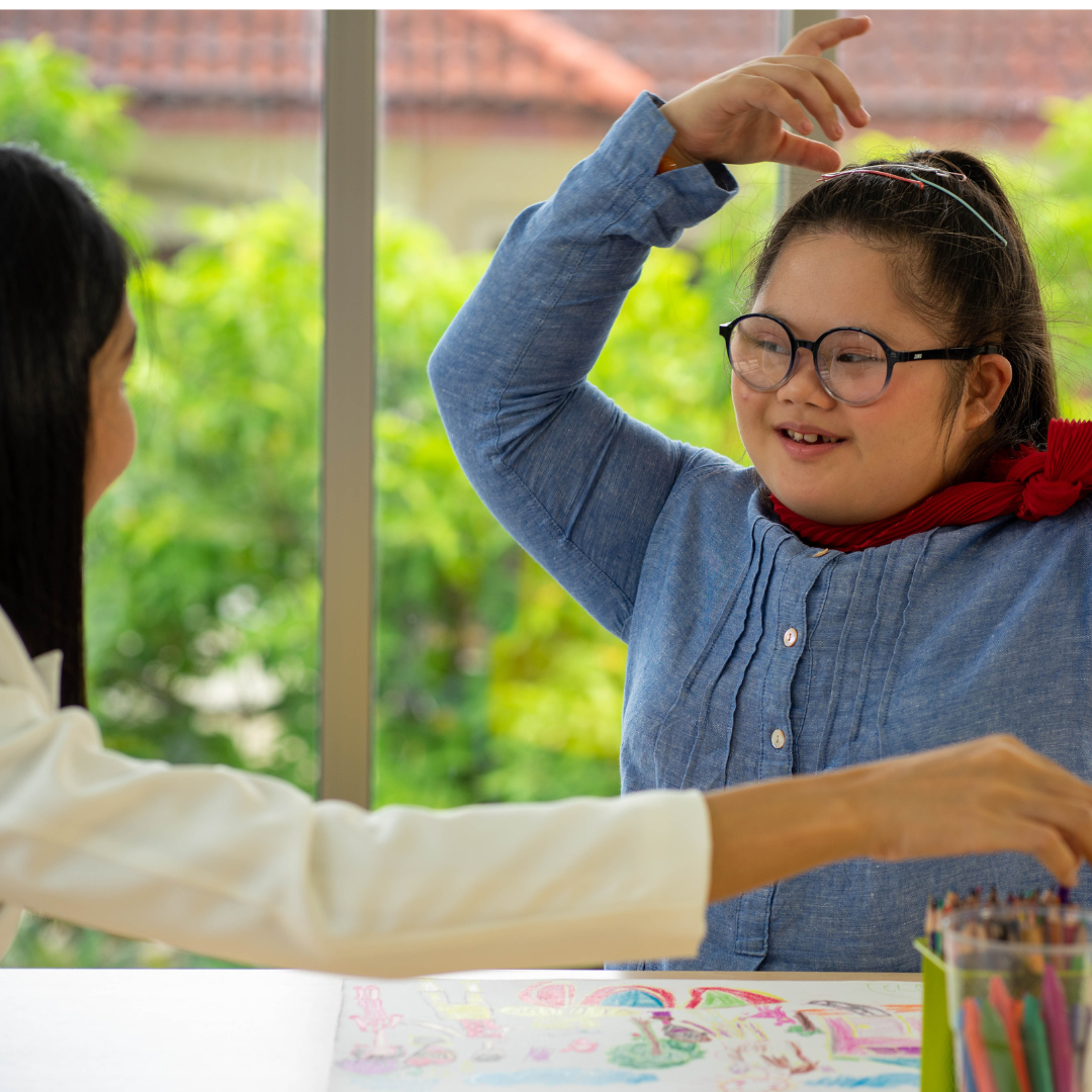 Girl sat at a drawing table wearing a cape smiling at an adult