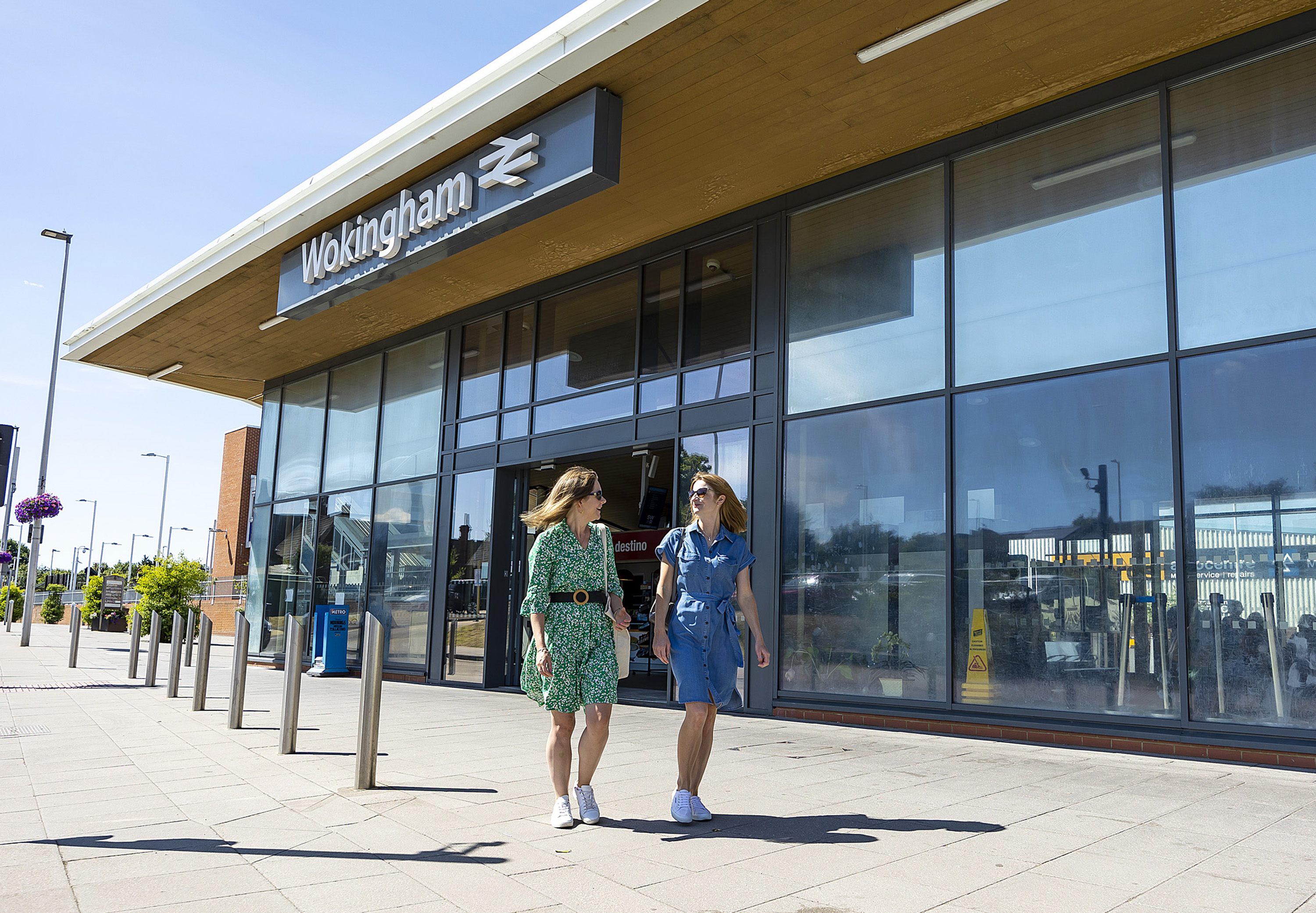Two women smile and chat with each other while walking past Wokingham train station on a sunny day